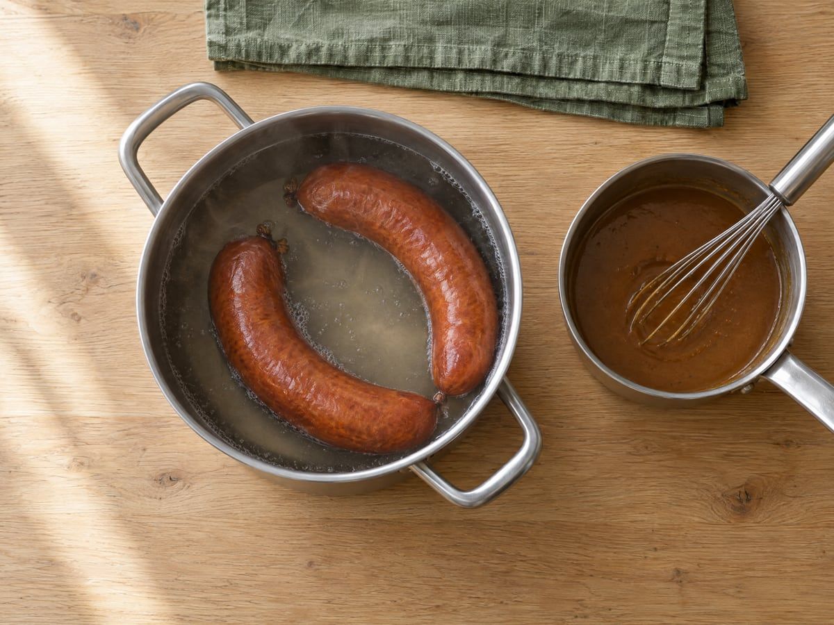 Twee rookworsten in een pan met heet water, naast een steelpannetje met jus