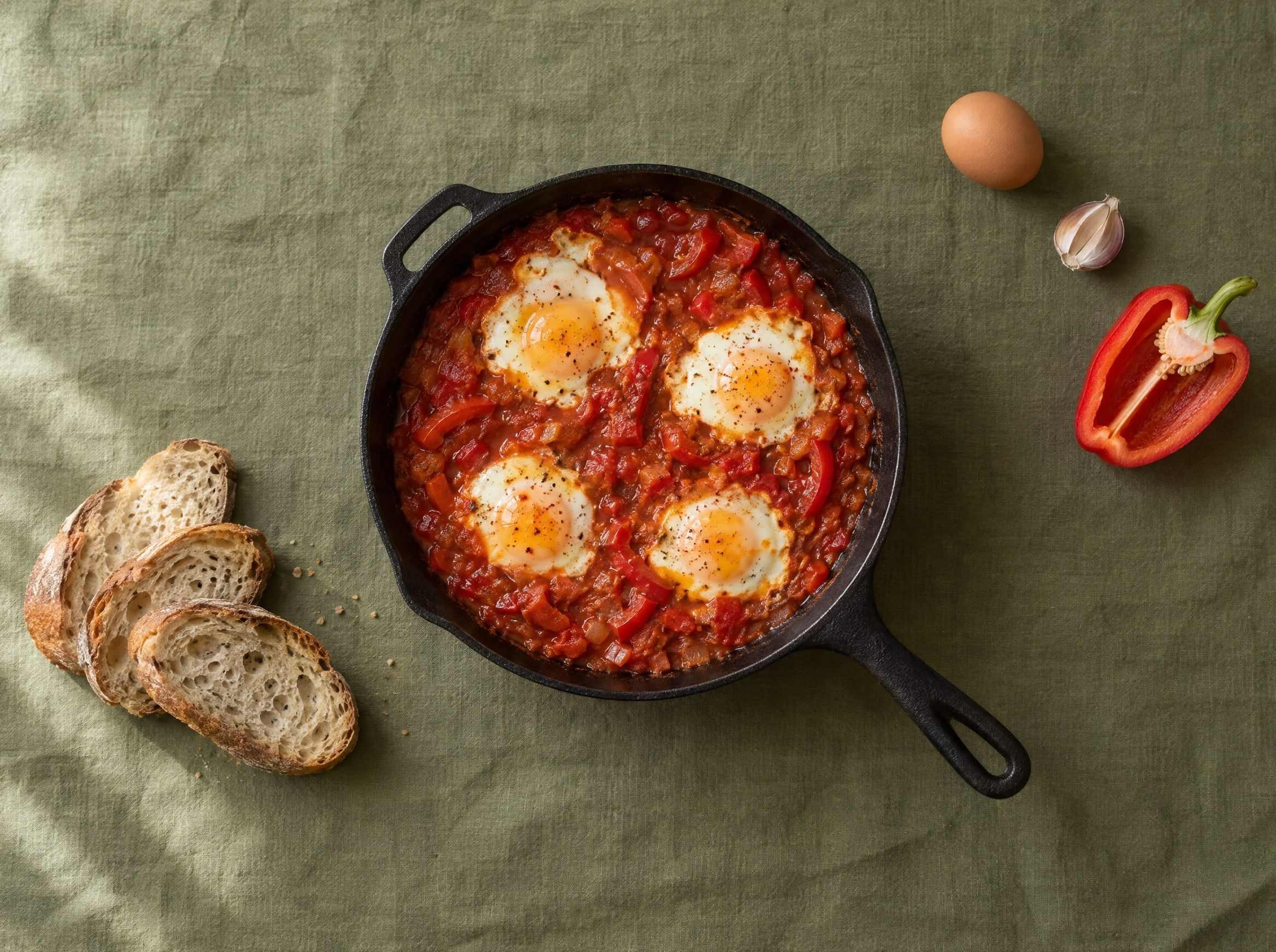 Shakshuka met vier eieren in tomaten-paprikasaus in een gietijzeren pan, met sneetjes brood, op een olijfgroen linnen kleed